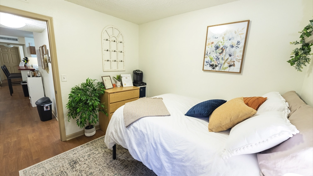 A bedroom featuring a white bedspread, colorful pillows, a dresser with framed photos, and a decorative plant. The view extends toward the kitchen area.
