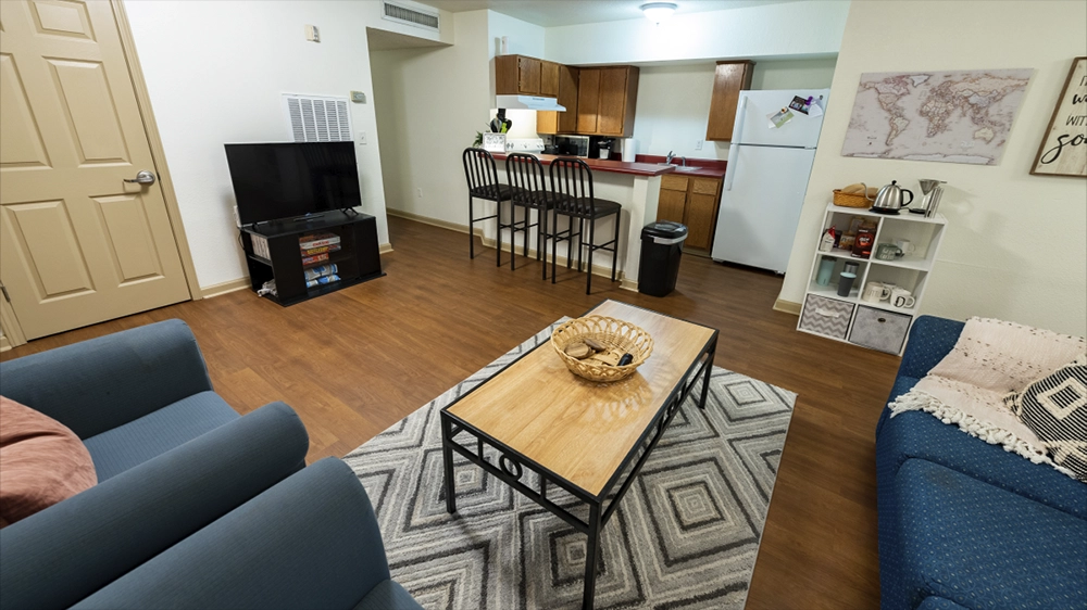 A furnished living area with two blue sofas, a wooden coffee table, and a TV stand. The open layout connects to a kitchen with wooden cabinets and a breakfast bar with stools.