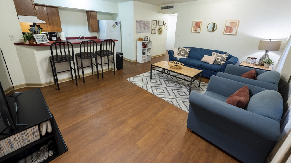 A living space with blue sofas, a patterned rug, and a wooden coffee table. The kitchen features a breakfast bar with stools and wooden cabinetry.