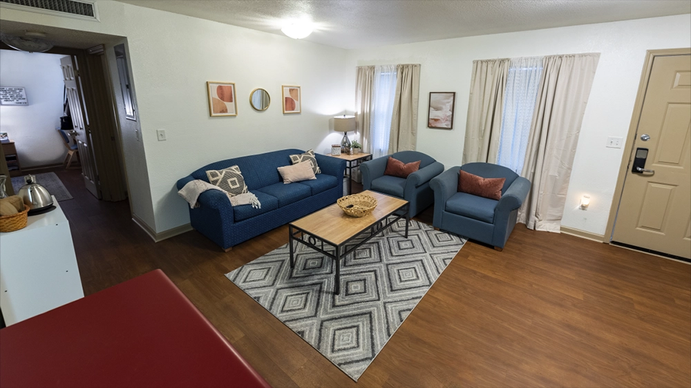 A living room with blue sofas, a patterned rug, and a coffee table, viewed from the kitchen area. Natural light filters through the windows.