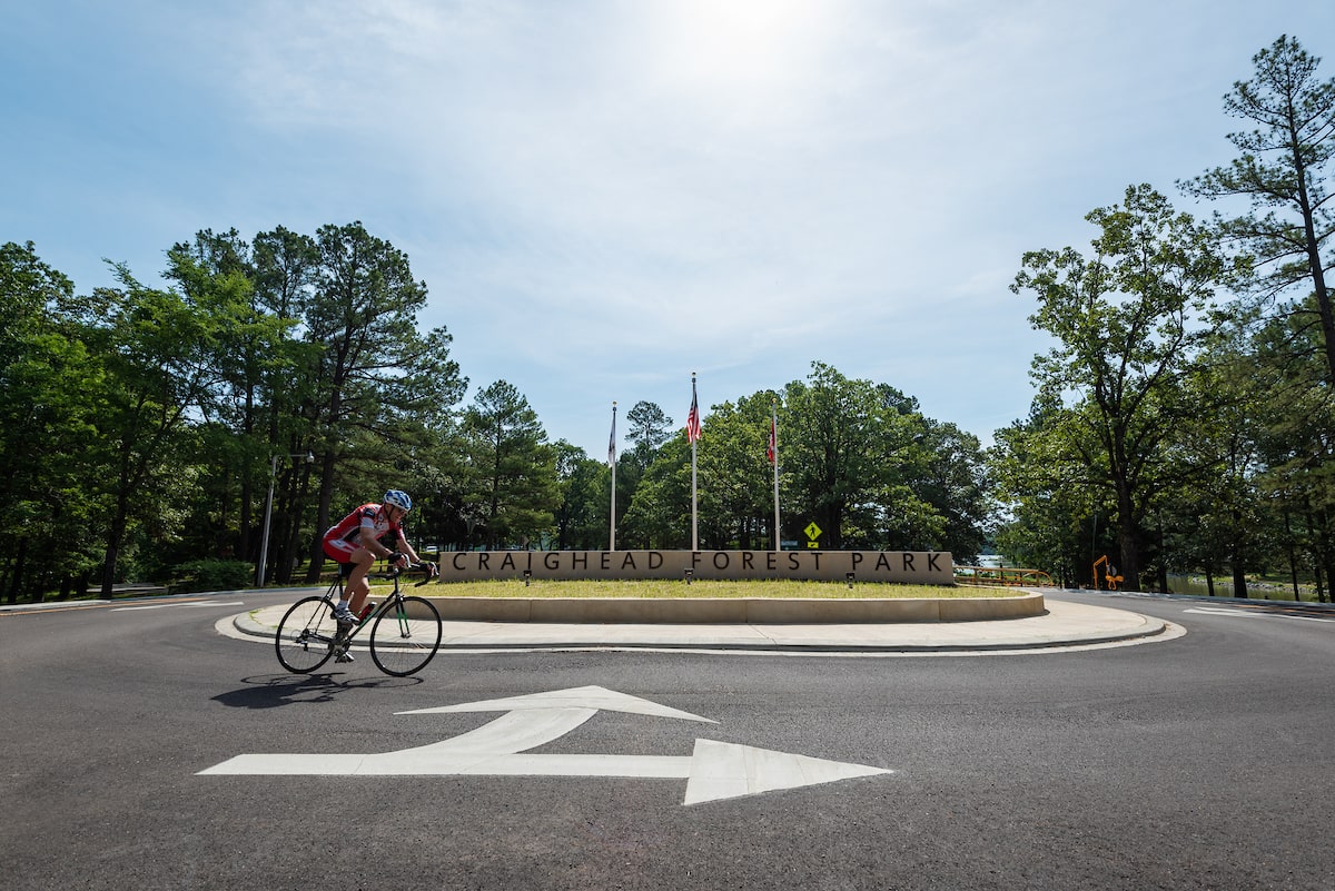 A cyclist rides past the Craighead Forest Park sign surrounded by trees and flags.