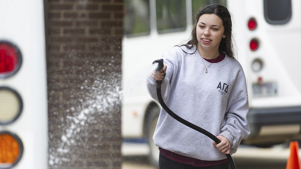 Individual wearing a gray Alpha Gamma Delta sweatshirt spraying water from a hose during a volunteer car wash.