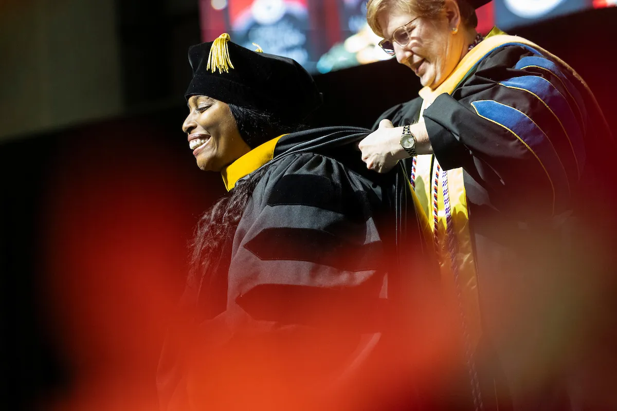 A graduate smiles while being hooded during a doctoral ceremony on stage.