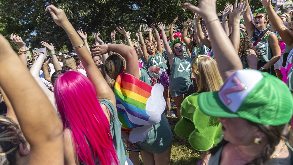 Crowd of people wearing green shirts and colorful accessories raising their hands outdoors during Bid Day festivities.