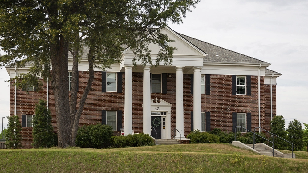 Two-story red brick sorority house featuring tall white columns and black shutters. A large tree partially frames the front view. The entrance has steps leading to a white door beneath a triangular pediment.