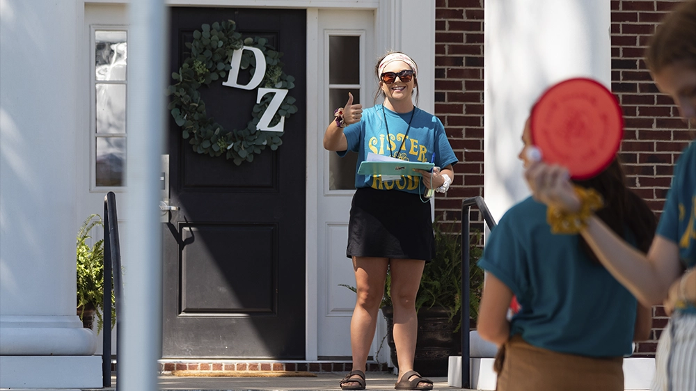 Individual standing in front of a black door decorated with a wreath and “DZ” letters, wearing a teal shirt that says “Sisterhood.”