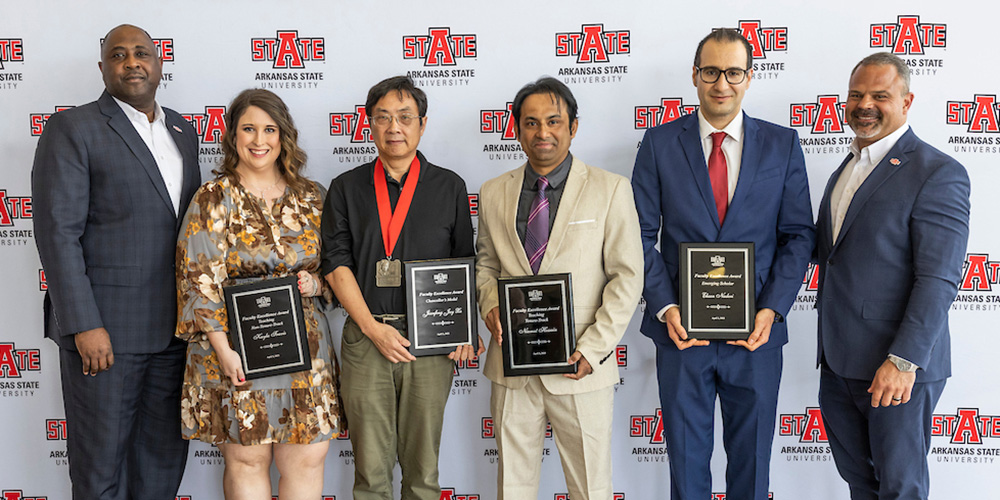 Group of faculty member award winners pose holding plaques in front of A-State backdrop during honors event.
