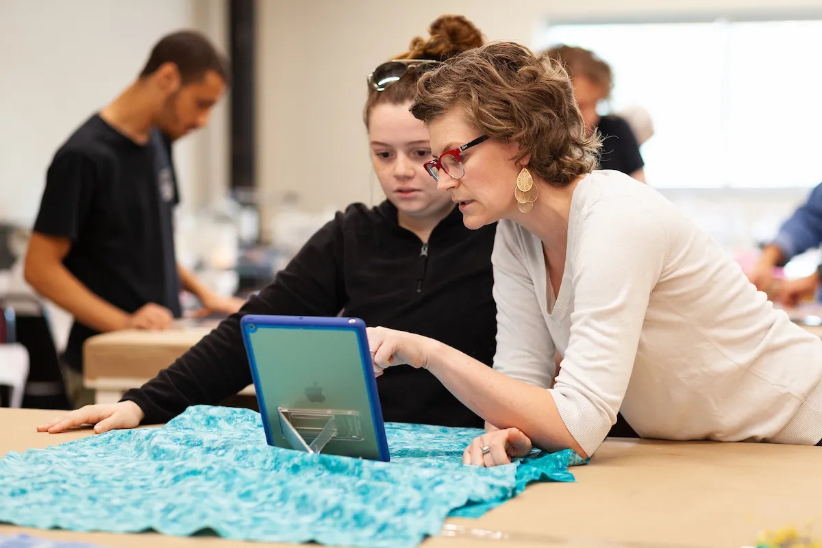 Instructor guides student with digital tablet while working on fabric design.