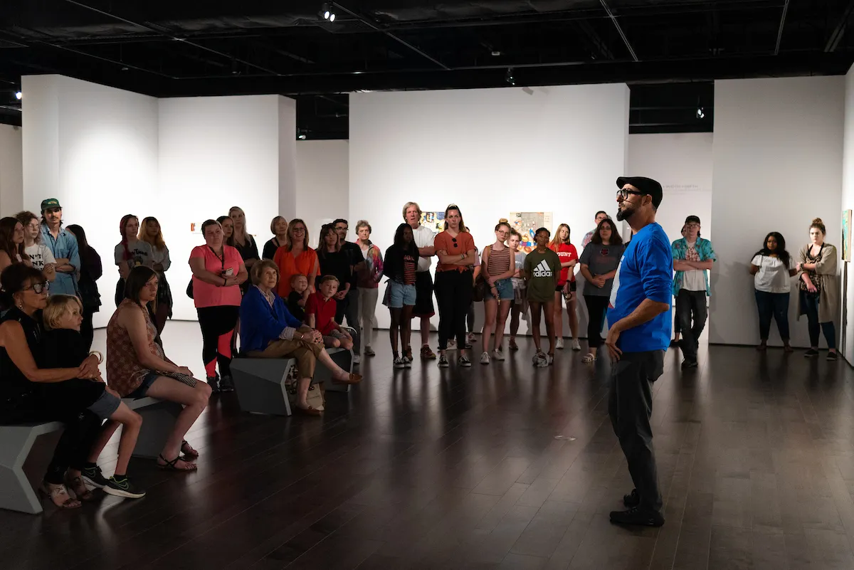 Group listens to artist speaking inside A-State art gallery during tour or event.