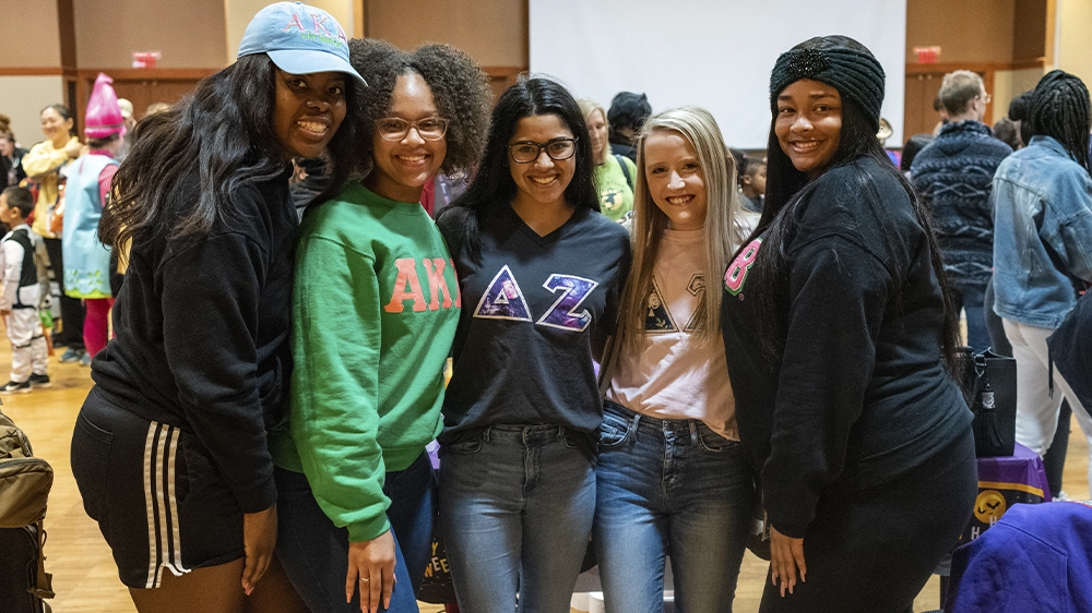 Group of individuals indoors wearing Greek-letter shirts posing together at a Halloween-themed event.