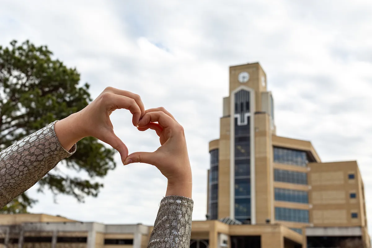 A student makes a heart gesture with their hands in front of the Dean B. Ellis Library