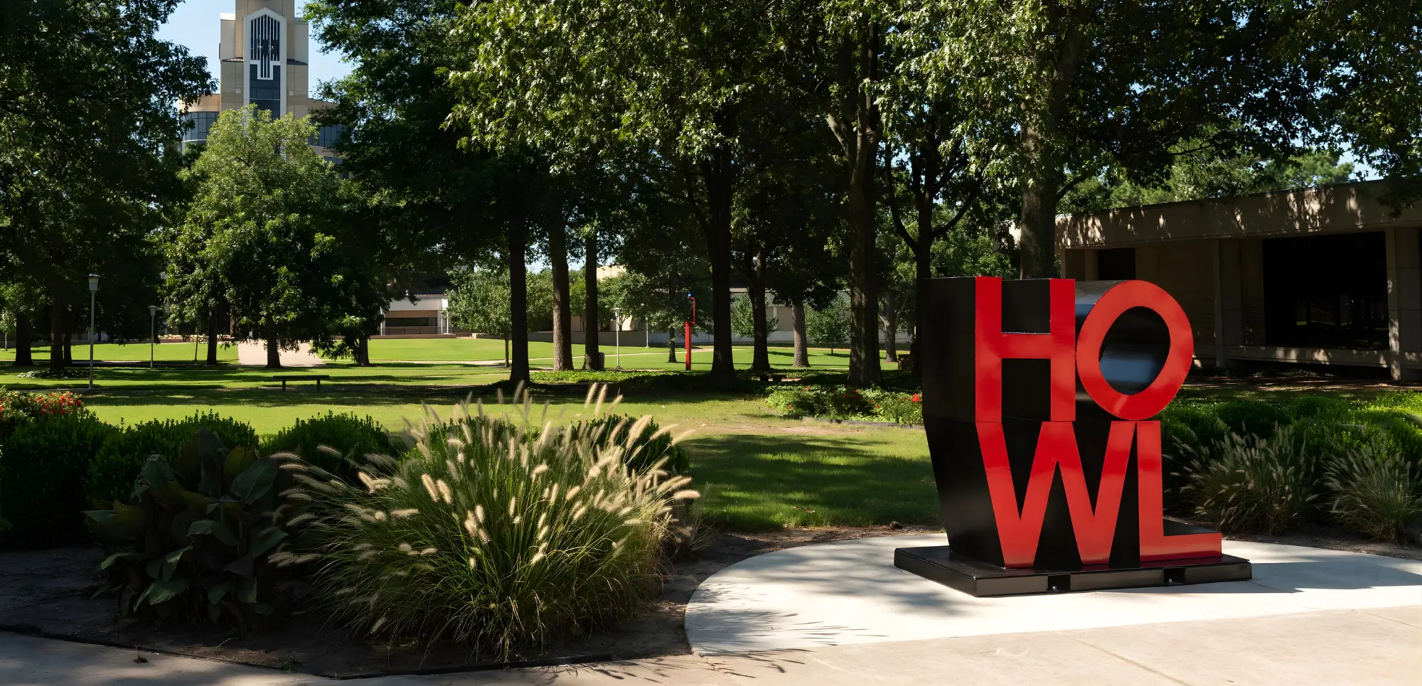 The red and black “HOWL” sculpture stands prominently on the Arkansas State University campus in Jonesboro, surrounded by trees and greenery. In the background, the Dean B. Ellis Library clock tower rises above the campus buildings on a bright, sunny day.
