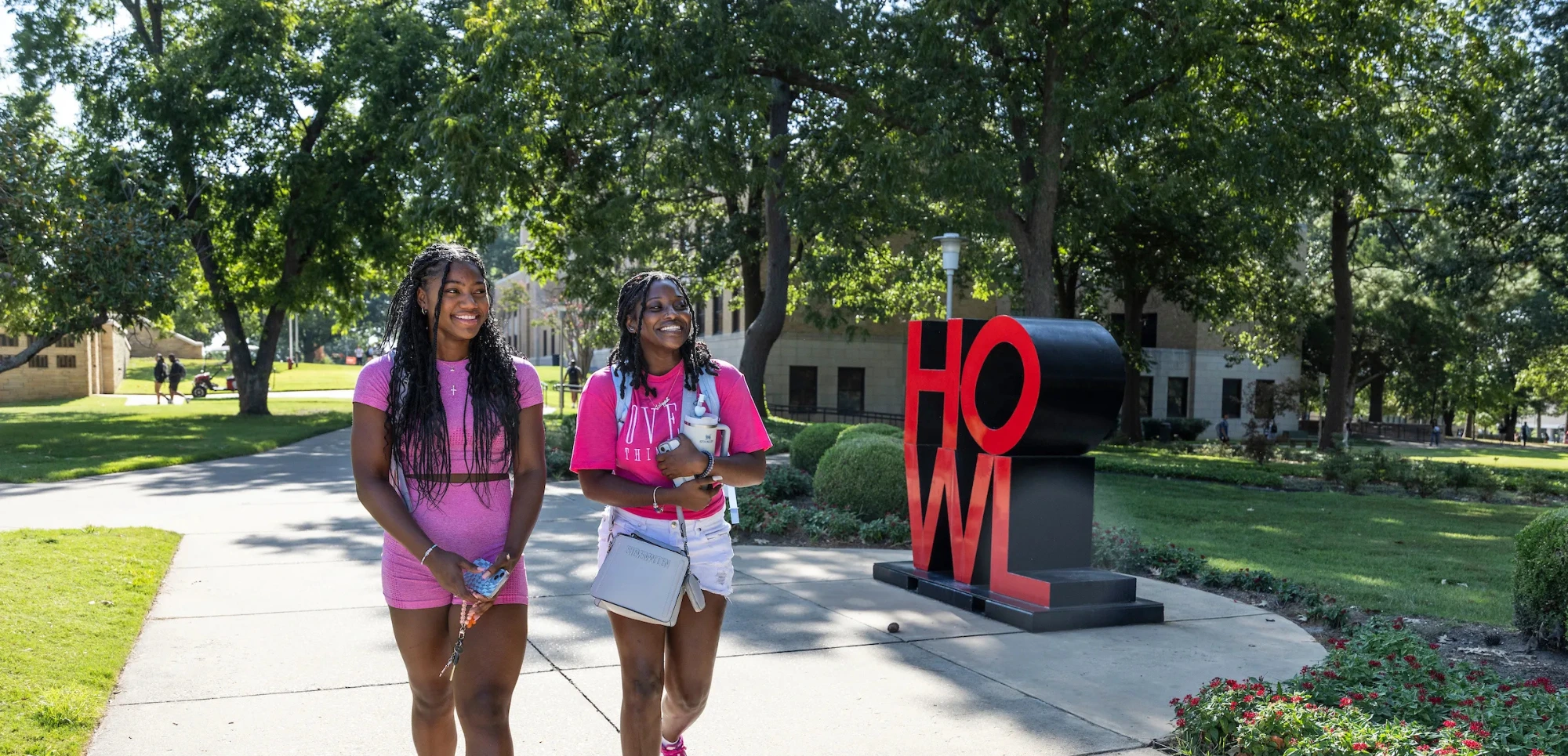 Two students in pink outfits walk past the HOWL sculpture on a sunny day at campus.