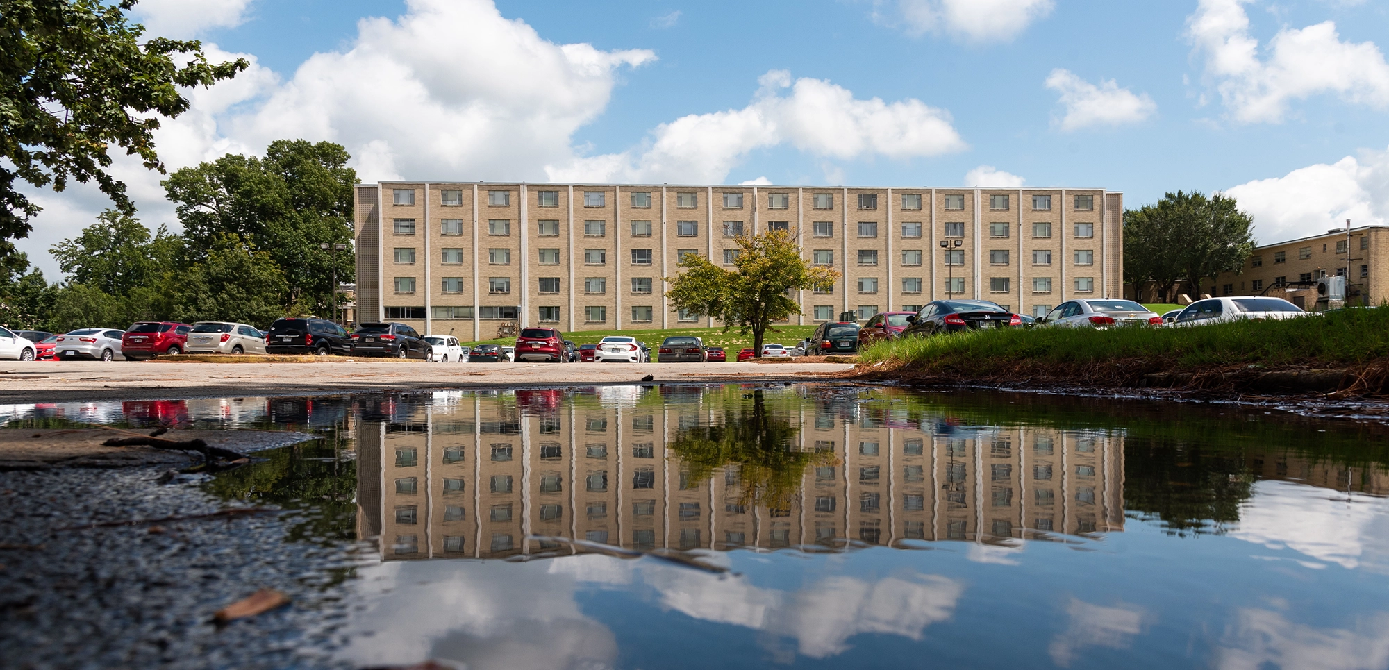 Exterior view of University Hall, a five-story brick residence building with rows of windows, cars parked in front, and its reflection visible in a large puddle under a partly cloudy sky.