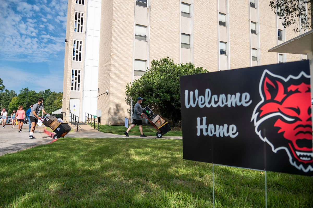 Sign that reads 'welcome home' in front of residence hall building with people taking items in and out a side door.