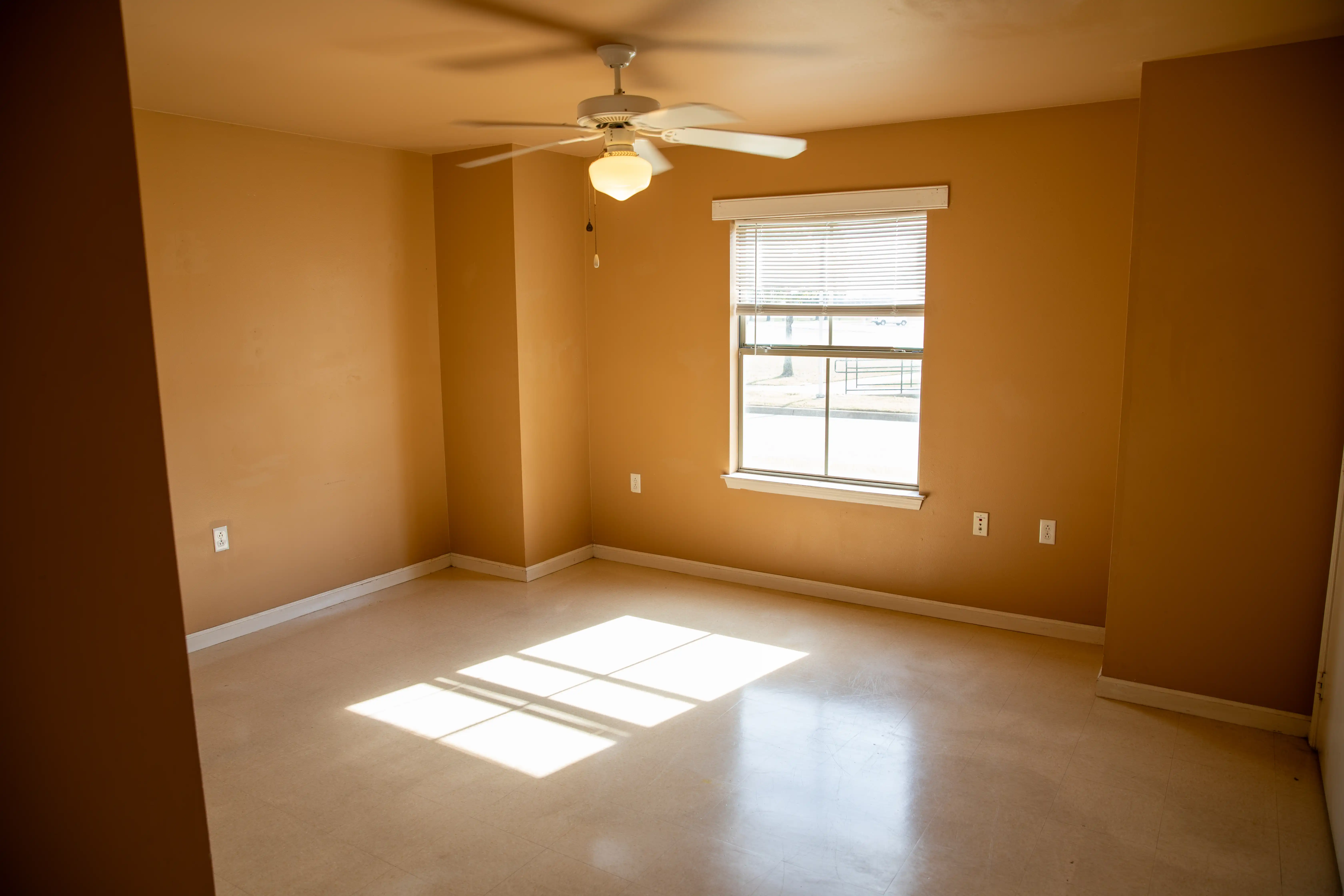 Empty bedroom with tan walls, ceiling fan, and window with blinds.