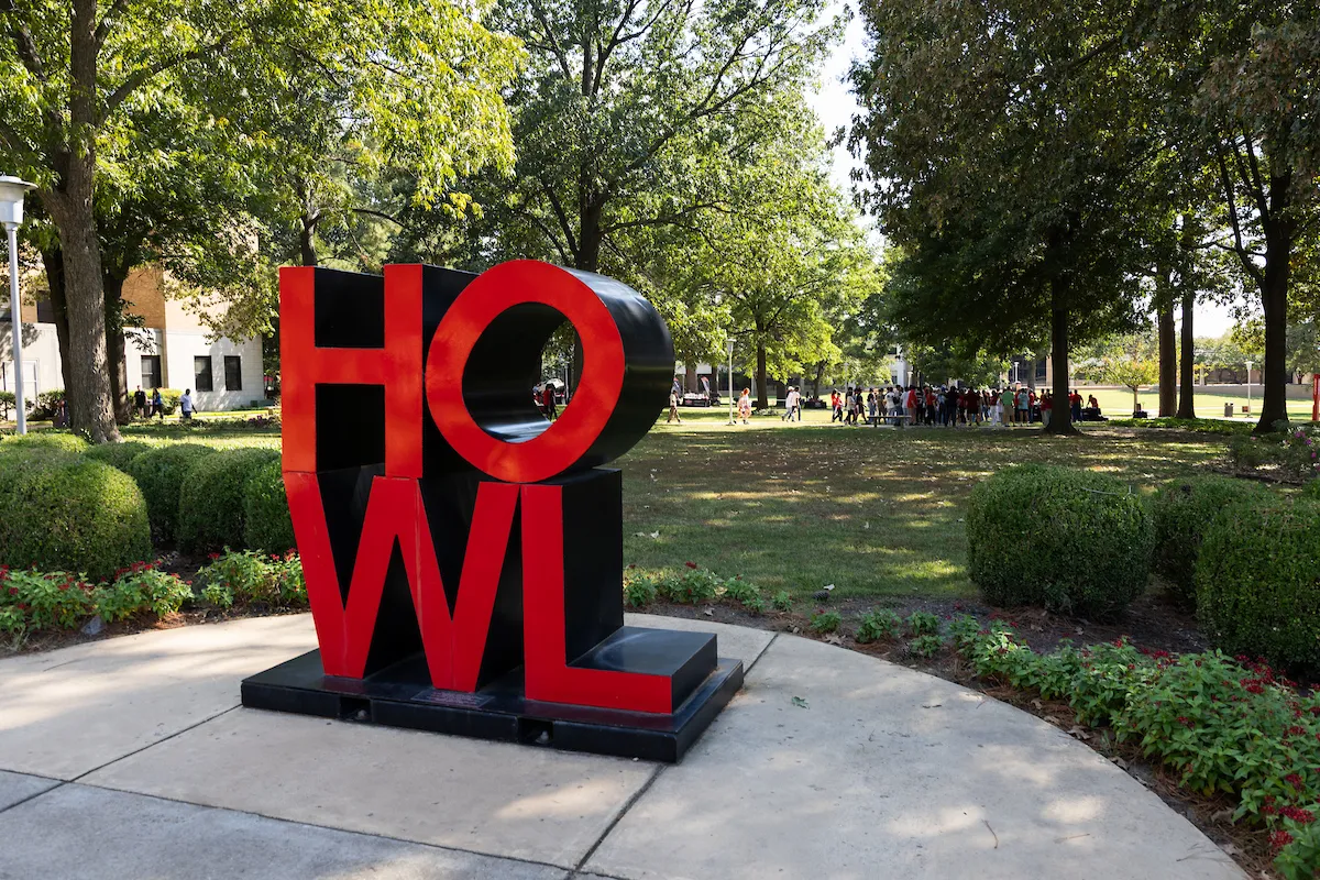 Bold red HOWL sculpture on A-State campus sidewalk surrounded by trees and trimmed hedges on a sunny day.