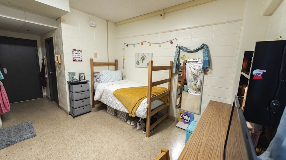 Single bed with white bedding and yellow throw, desk, and mirror in Kays Hall room.