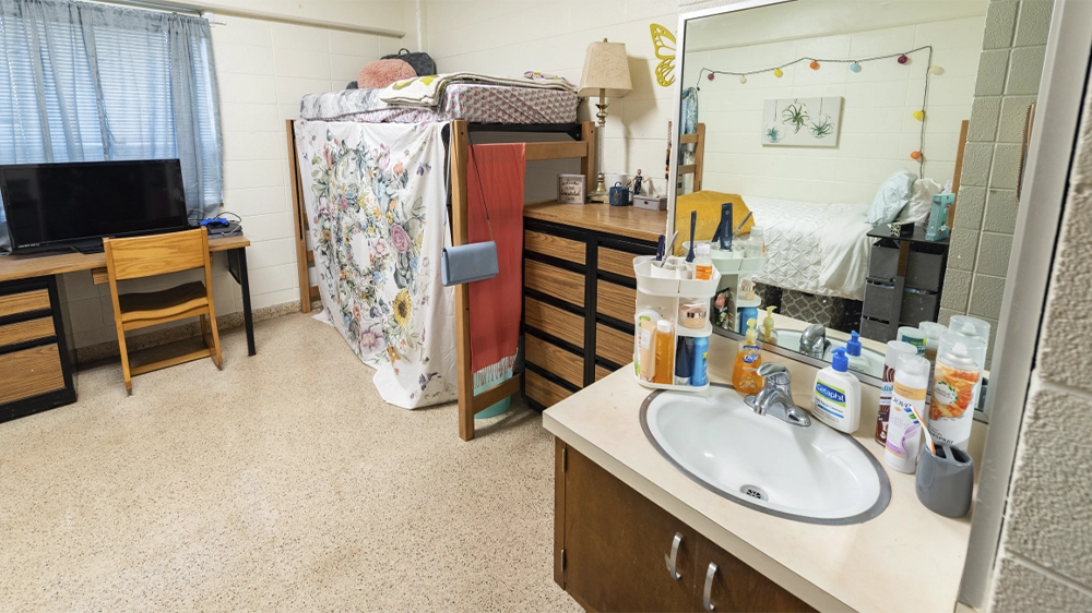 Kays Hall bedroom showing lofted bed, dresser, and sink with mirror in foreground.