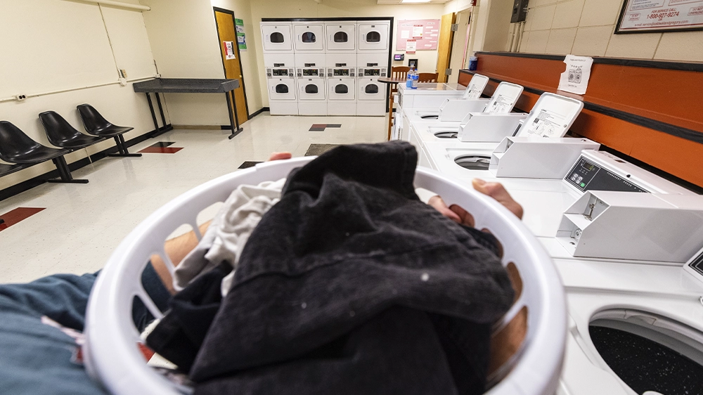 Laundry room with multiple washers and dryers, folding tables, and seating along the wall. A laundry basket filled with clothes is visible in the foreground.