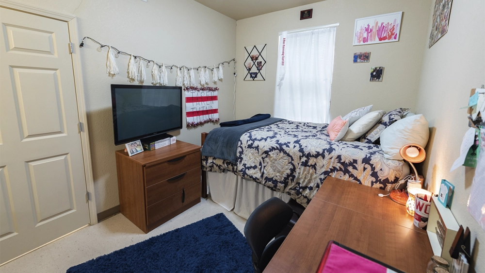 Bedroom with a twin bed featuring floral bedding, wooden dresser with TV, desk with chair, and door leading to entryway.