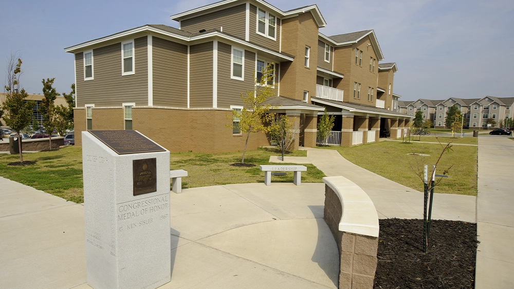 Exterior view of ROTC Living Learning Community building with tan brick and gray siding, featuring a landscaped walkway and a granite monument honoring G. Ken Sisler in the foreground.