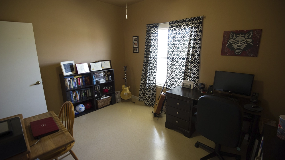 Bedroom with desk, bookshelf, guitar, and window with patterned curtains.