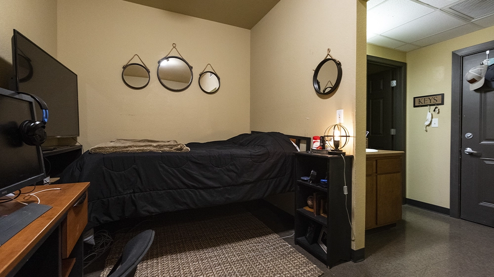 Bedroom with raised twin bed, black bedding, desk with computer setup, and three decorative mirrors on the wall.