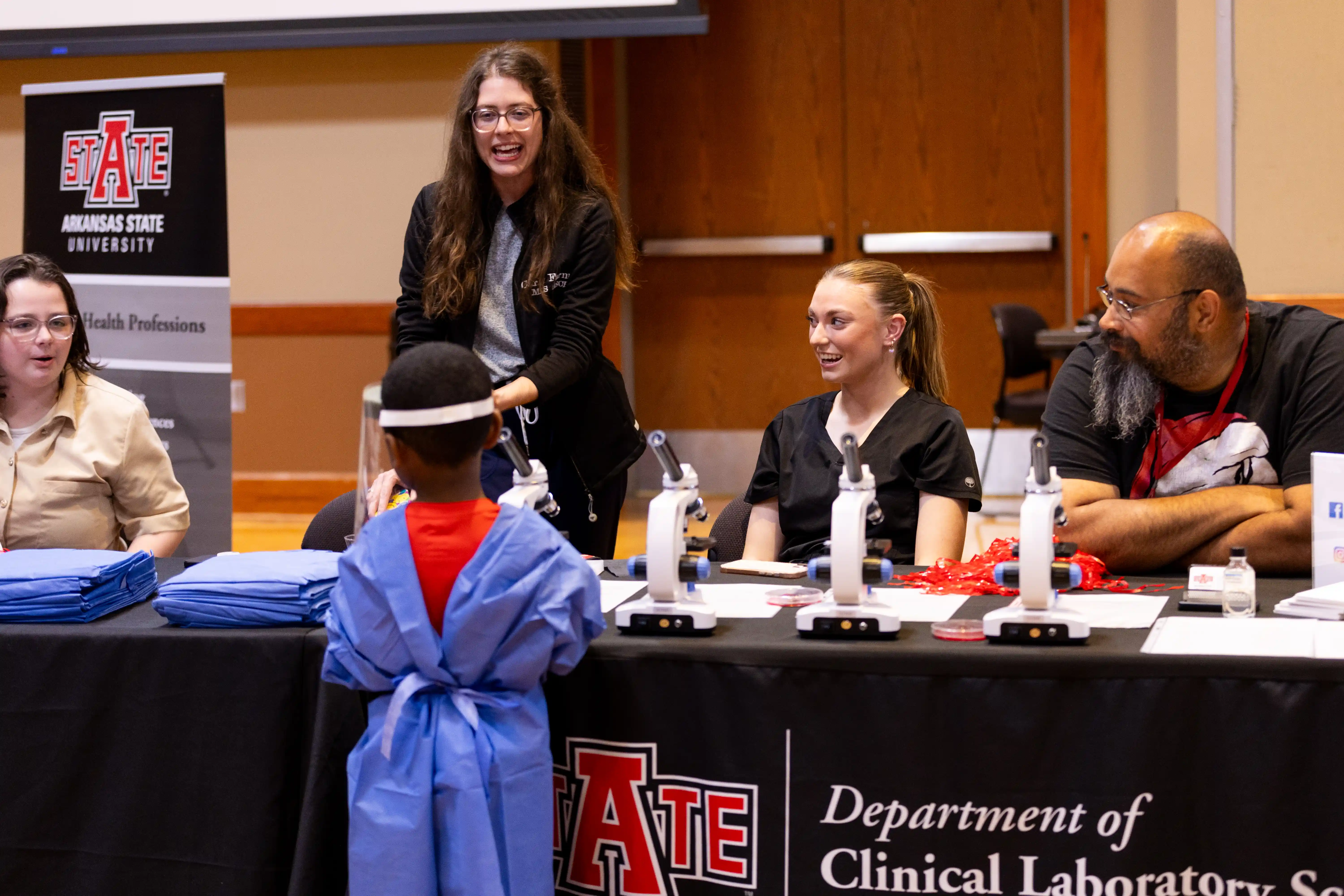 Clinical lab science booth at a fair with a kid in surgical scrubs and face mask walks around looking at equiptment.