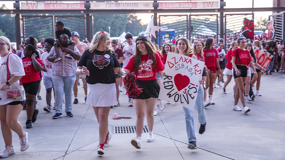 Crowd walking through stadium gates wearing red and carrying signs supporting Red Wolves and Greek organizations.