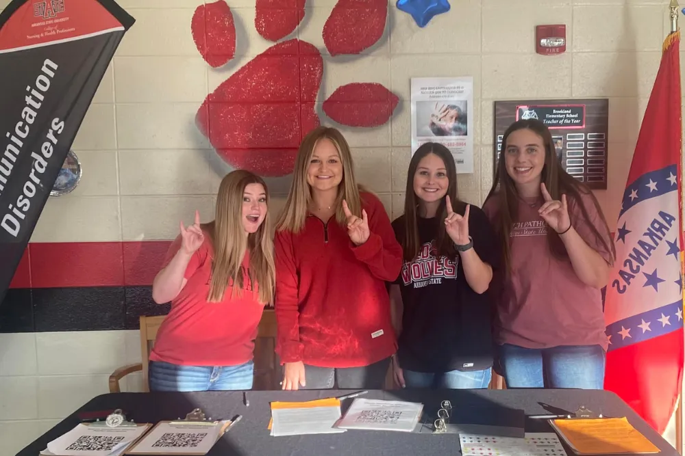 Communication Disorders students at an event sitting at the communication disorders booth while they give a wolves up sign.