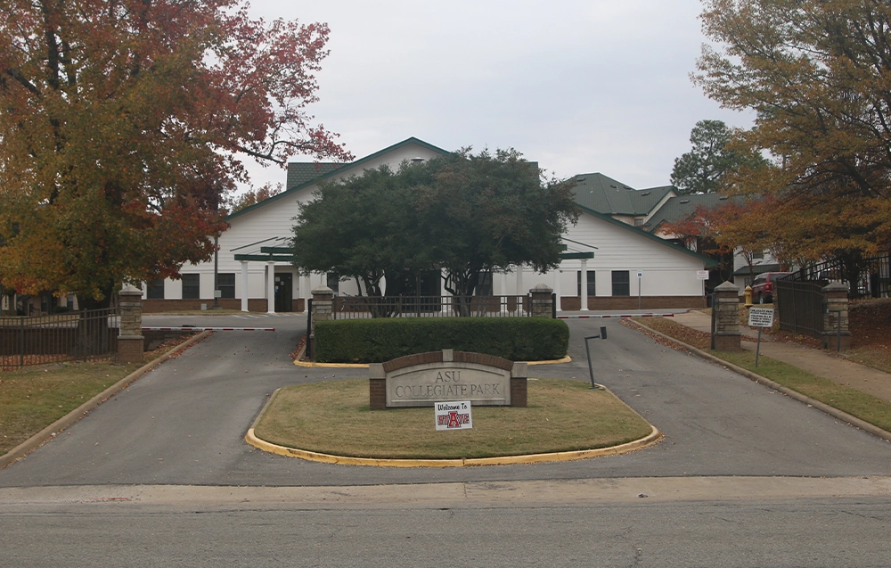 Entrance view of ASU Collegiate Park featuring a wide driveway flanked by stone pillars and fencing, a landscaped median with a sign reading “ASU Collegiate Park,” and a large white building with green roof in the background surrounded by autumn trees.