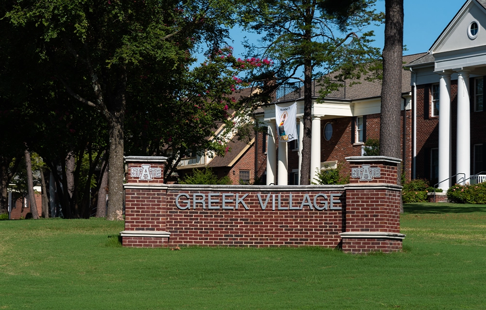 Brick monument sign reading “Greek Village” with Arkansas State University logos on each side, set on a manicured lawn. Red brick sorority houses with white columns and trees are visible in the background.