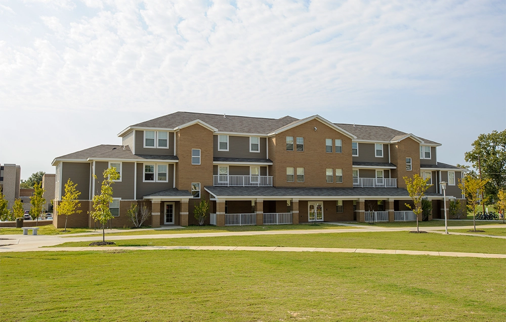 Exterior view of a three-story apartment-style building with tan brick and gray siding, featuring multiple balconies and surrounded by green lawns under a partly cloudy sky.