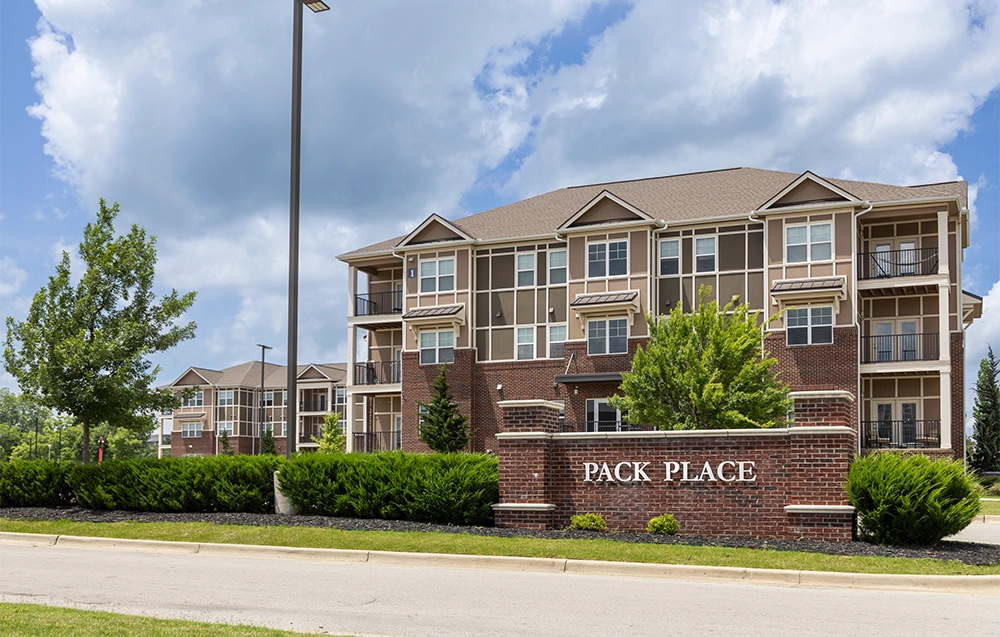 Exterior view of Pack Place apartments featuring two three-story brick buildings with large screened balconies, landscaped greenery, and a sign reading “Pack Place” in front.
