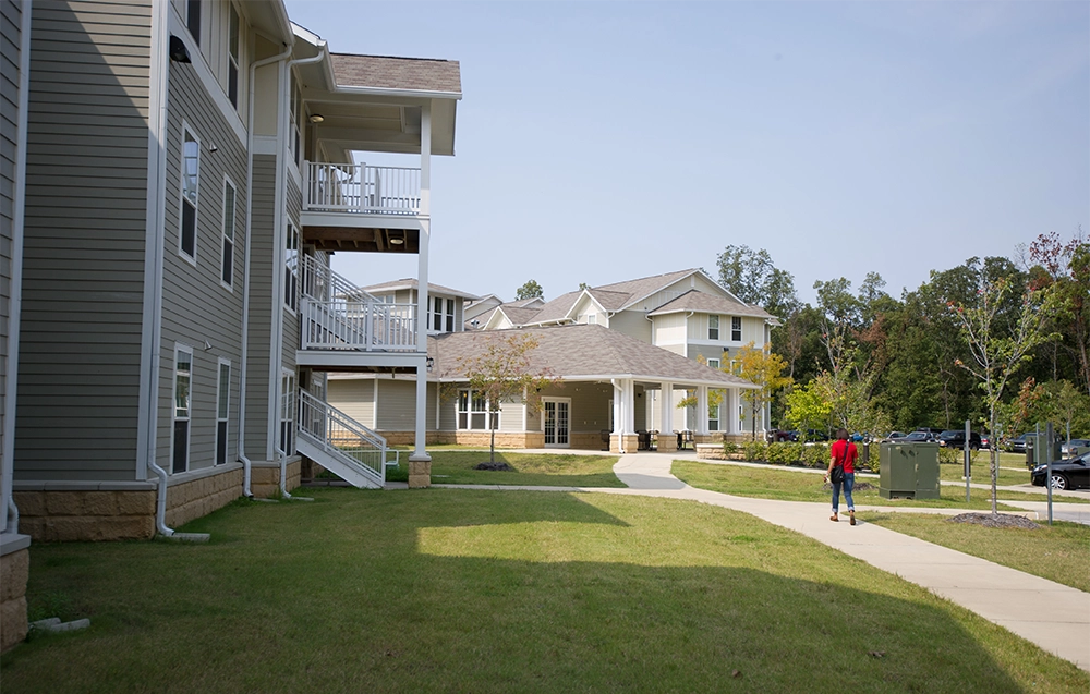 Side view of Red Wolf Den apartment buildings with light gray siding and white trim, connected by paved walkways and landscaped green lawns. A person is walking along the sidewalk toward the commons building in the background, surrounded by trees and parked cars.