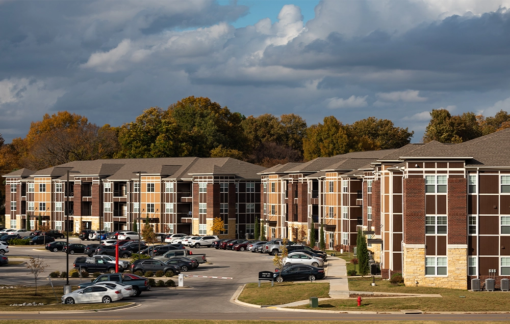 Wide view of The Circle apartment buildings with multiple stories, brick exteriors, and large windows, with a parking lot full of cars in the foreground and trees in the background.