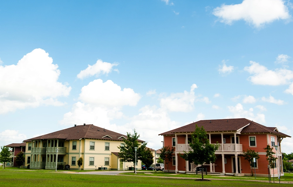 Two apartment buildings at The Village with light yellow and terracotta exteriors, white trim, and covered balconies, surrounded by green lawns and small trees under a bright blue sky with scattered clouds.