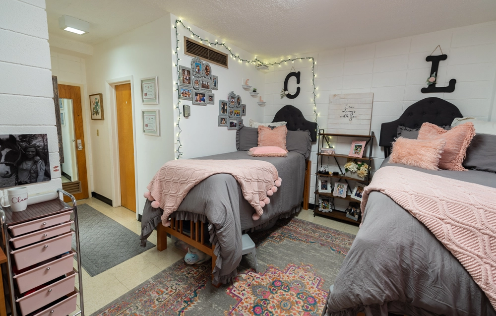 University Hall double bedroom decorated with gray bedding and pink accents. Two twin beds are arranged side by side with matching pillows and throws, a colorful area rug on the floor, and string lights framing the wall above photo collages and decorative letters.