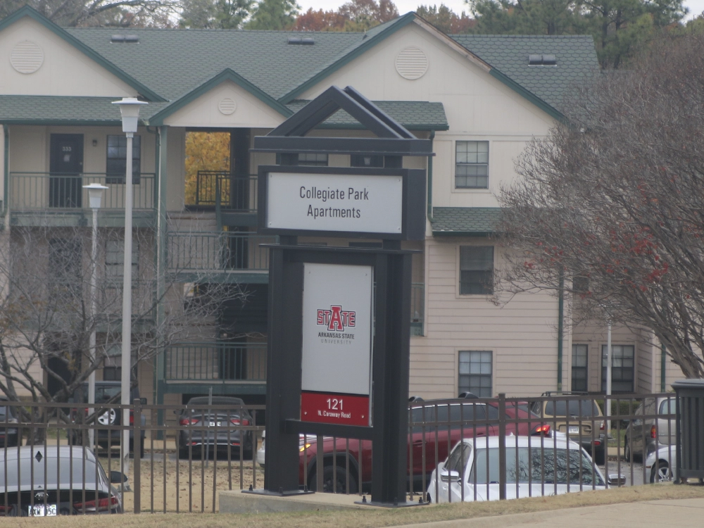 Sign for Collegiate Park Apartments with Arkansas State University branding and address “121 N. Caraway Road,” positioned in front of a multi-story apartment building with green roofs, balconies, and parked cars visible behind a black metal fence.