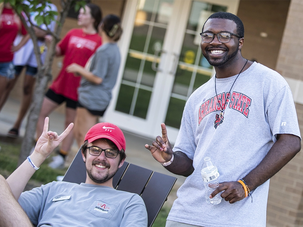 Two students in Arkansas State apparel sitting and standing outside a campus building, both making the Wolves Up hand sign, with other students talking in the background near a glass door entrance.