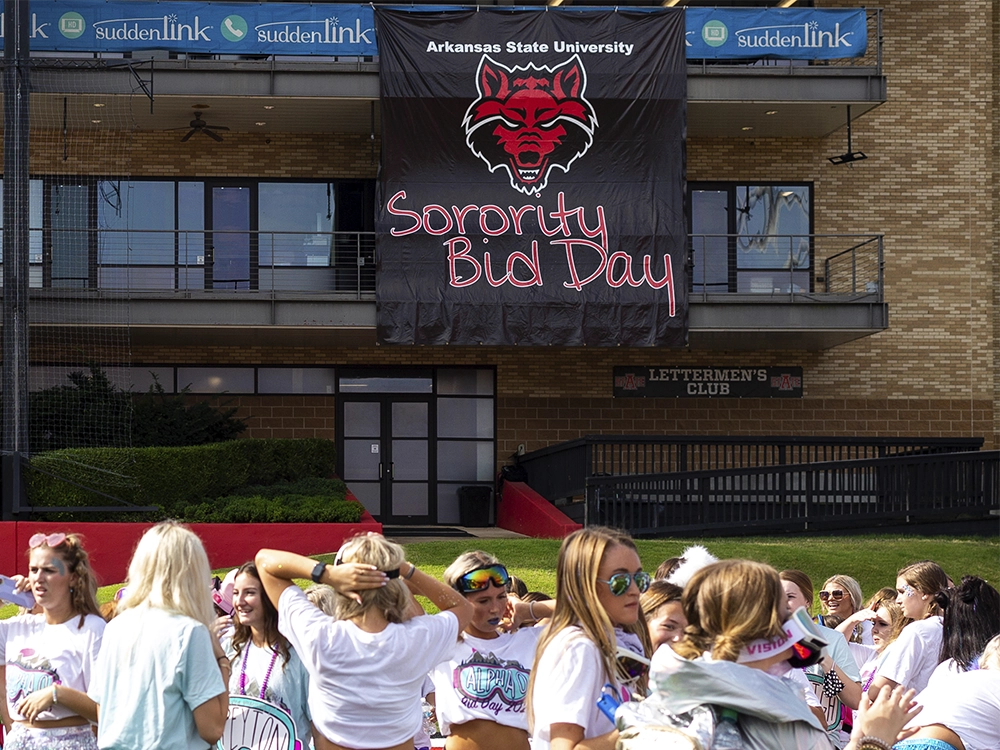 Large black banner with the Arkansas State University Red Wolves logo and the words “Sorority Bid Day” in pink letters hanging on a brick building. A group of people wearing matching shirts stands in front of the building.