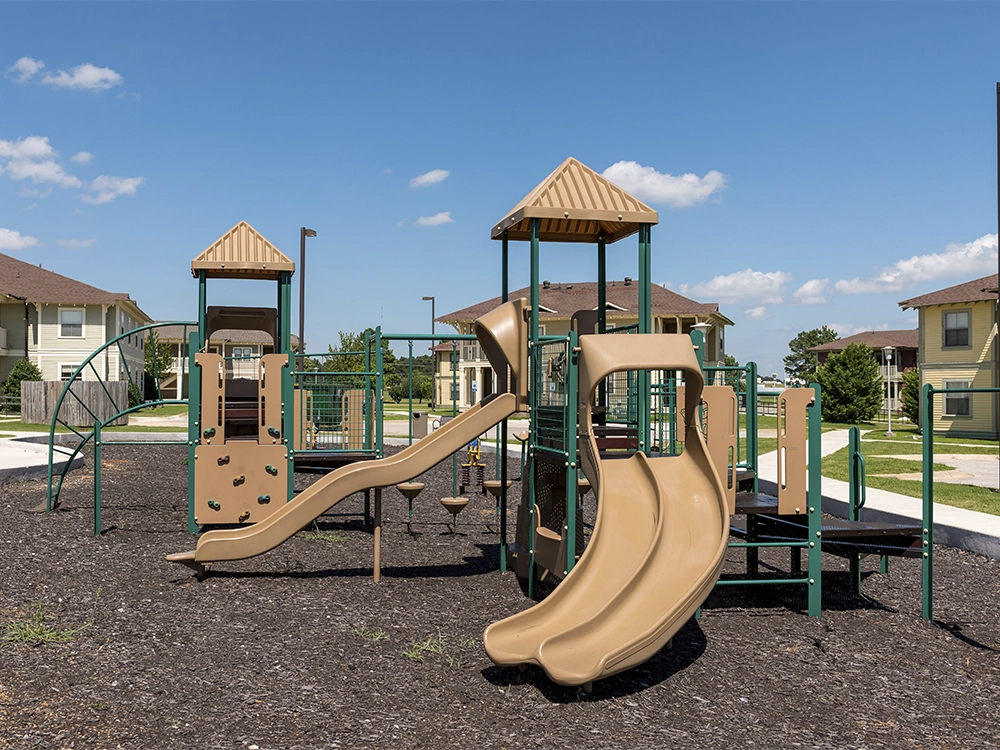 Outdoor playground at The Village featuring tan and green play structures with slides, climbing walls, and swings, surrounded by mulch and green lawns with apartment buildings in the background under a sunny blue sky.