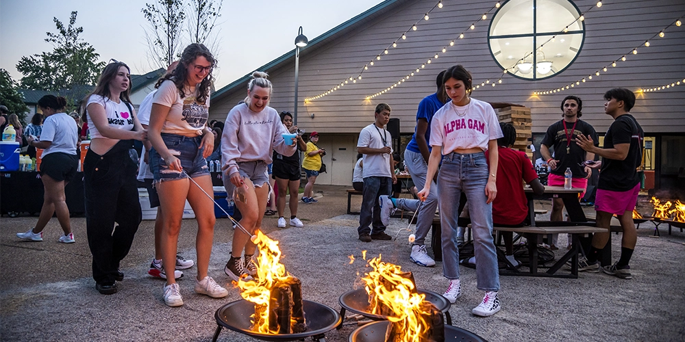 Outdoor social event at Collegiate Park with groups of people gathered around small fire pits roasting food, string lights overhead, and a building with a large circular window in the background creating a festive atmosphere.