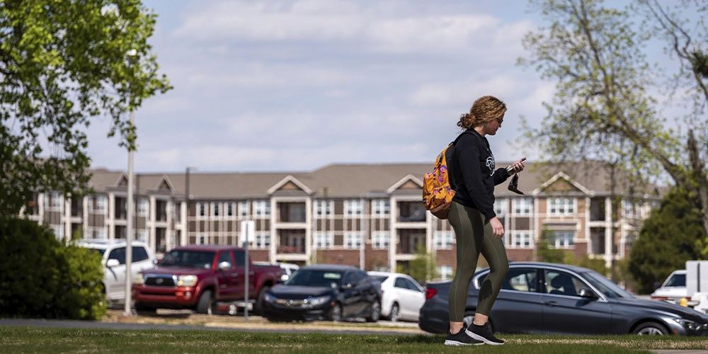 Student walking across a grassy area with cars parked nearby and Pack Place apartment buildings visible in the background under a partly cloudy sky.
