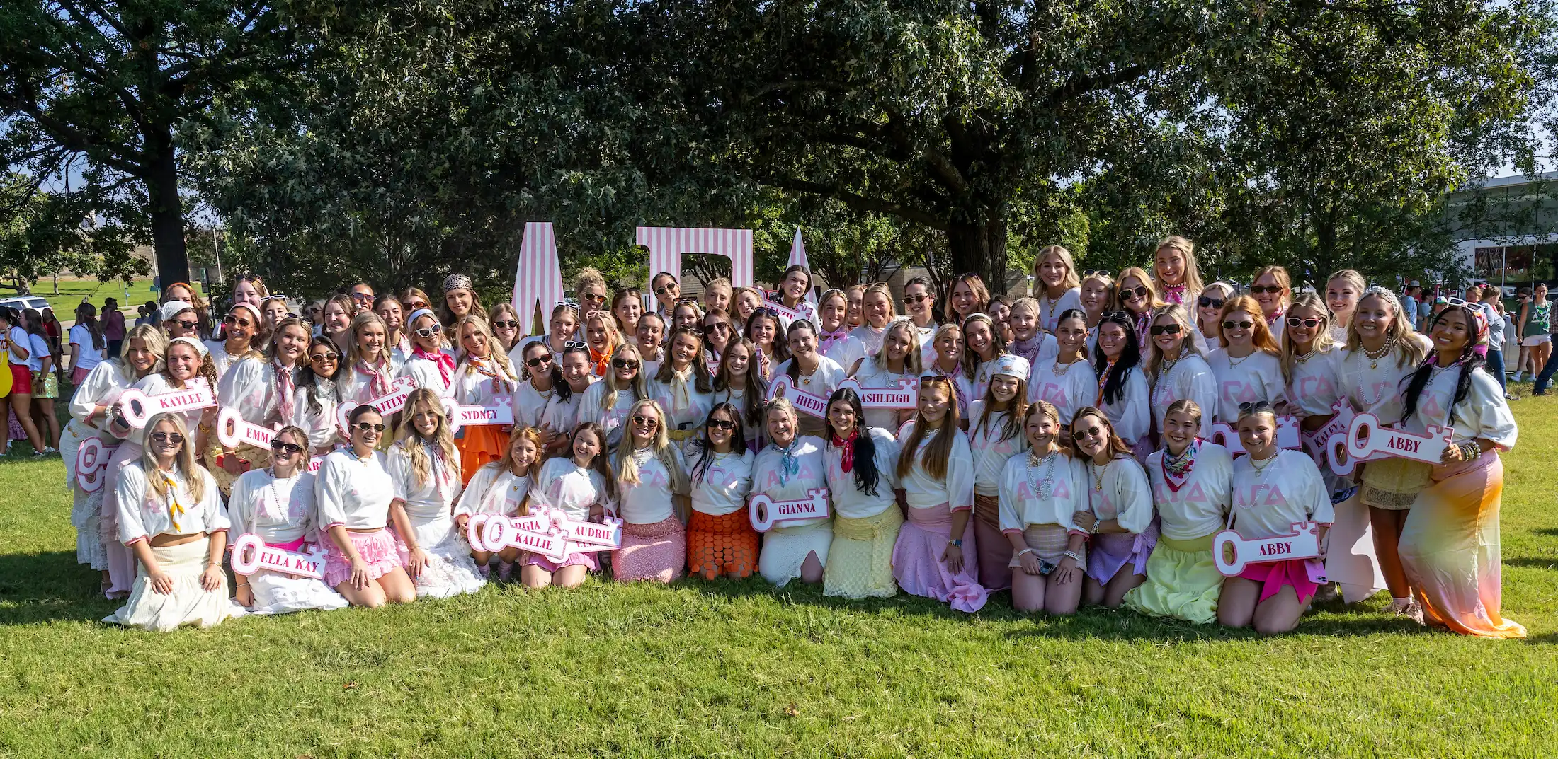 Wide outdoor shot of a large group of people standing and knealing on a grassy lawn holding colorful signs and Greek letters during Sorority Bid Day. 