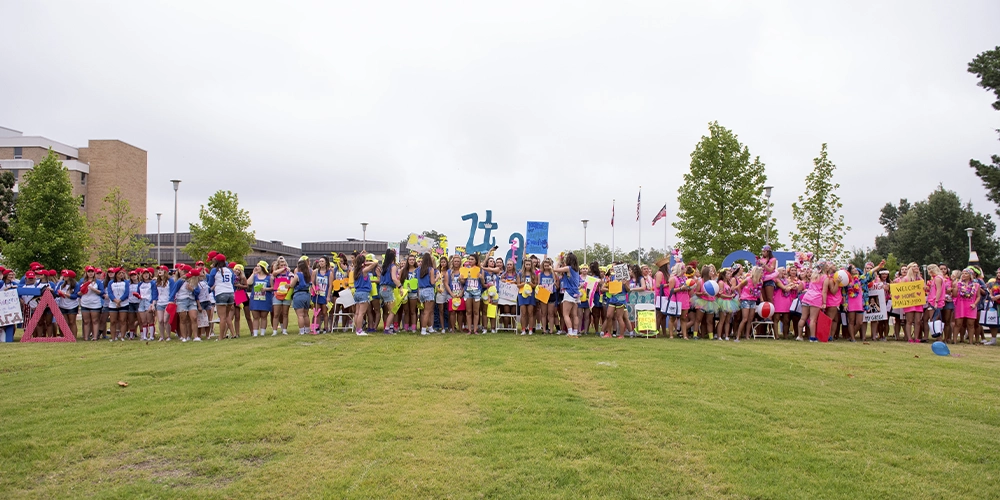 Wide outdoor shot of a large group of people standing on a grassy lawn holding colorful signs, balloons, and Greek letters during Sorority Bid Day. Multiple sorority groups are visible in bright shirts, with campus buildings and flagpoles in the background.