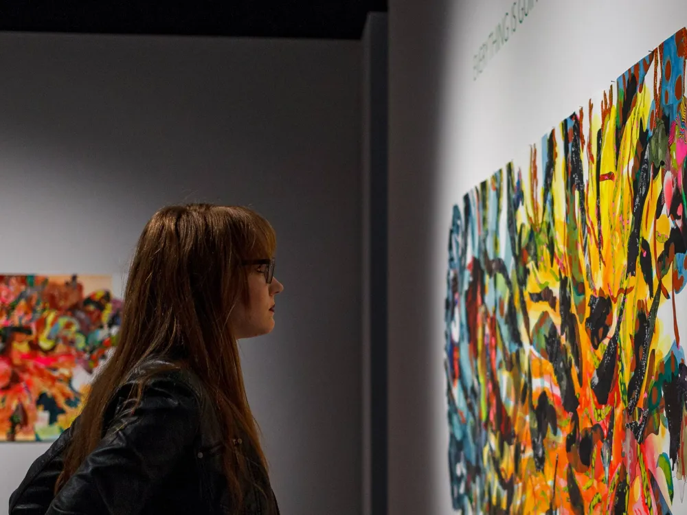 A person standing in an art gallery closely observing a large, colorful abstract painting on a white wall, with another vibrant artwork visible in the background.