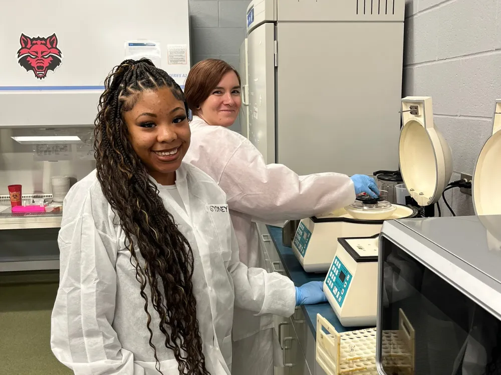 Two students in medical lab science class standing at a centrifuge smiling at the camera with a redwolf logo on the cabinet behind them.