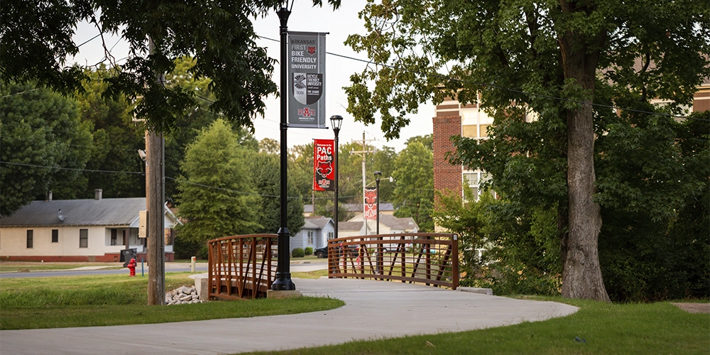 Curved sidewalk leading to a wooden pedestrian bridge near The Circle apartments, lined with trees and campus banners on lamp posts.